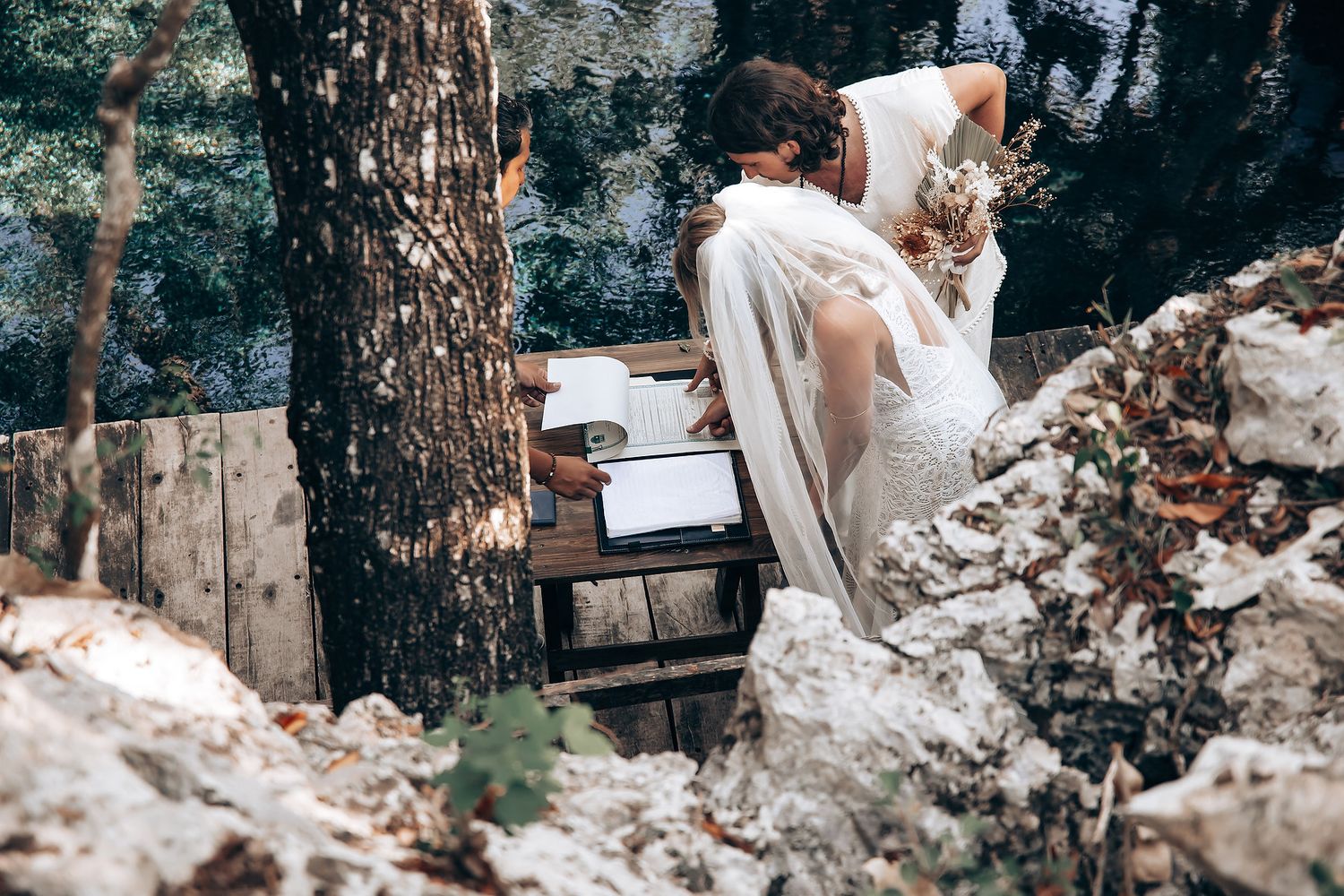 A couple in white attire shares a romantic moment in a rustic outdoor setting among trees and rocks.