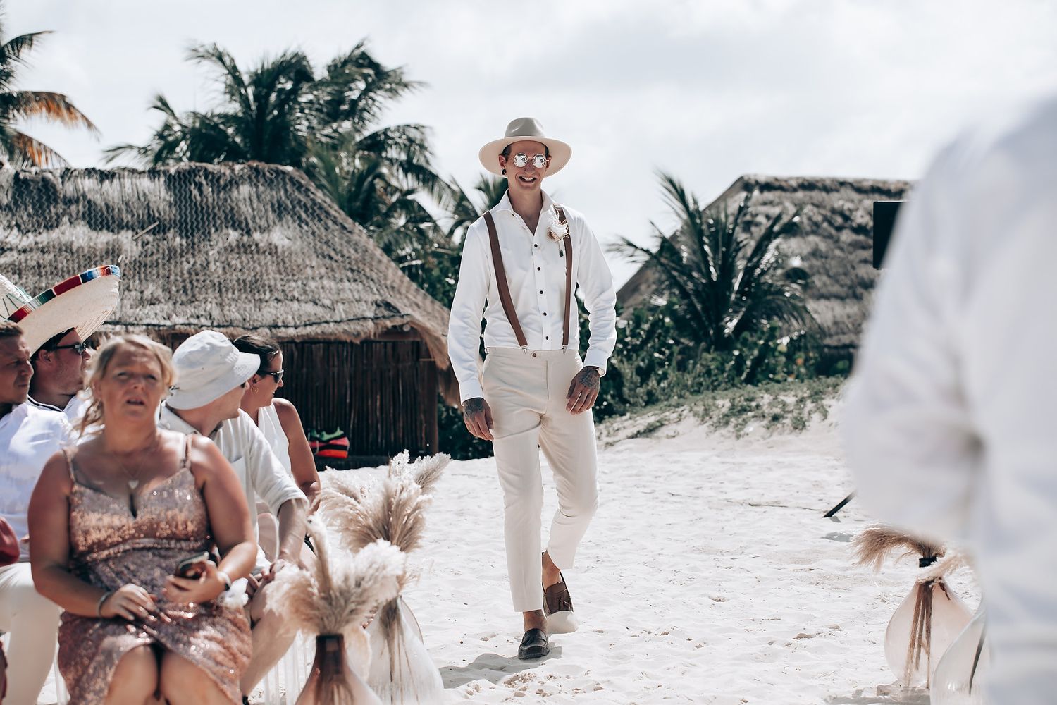 Beachside wedding celebration with guests in white attire gathered on sandy shores under palm trees.