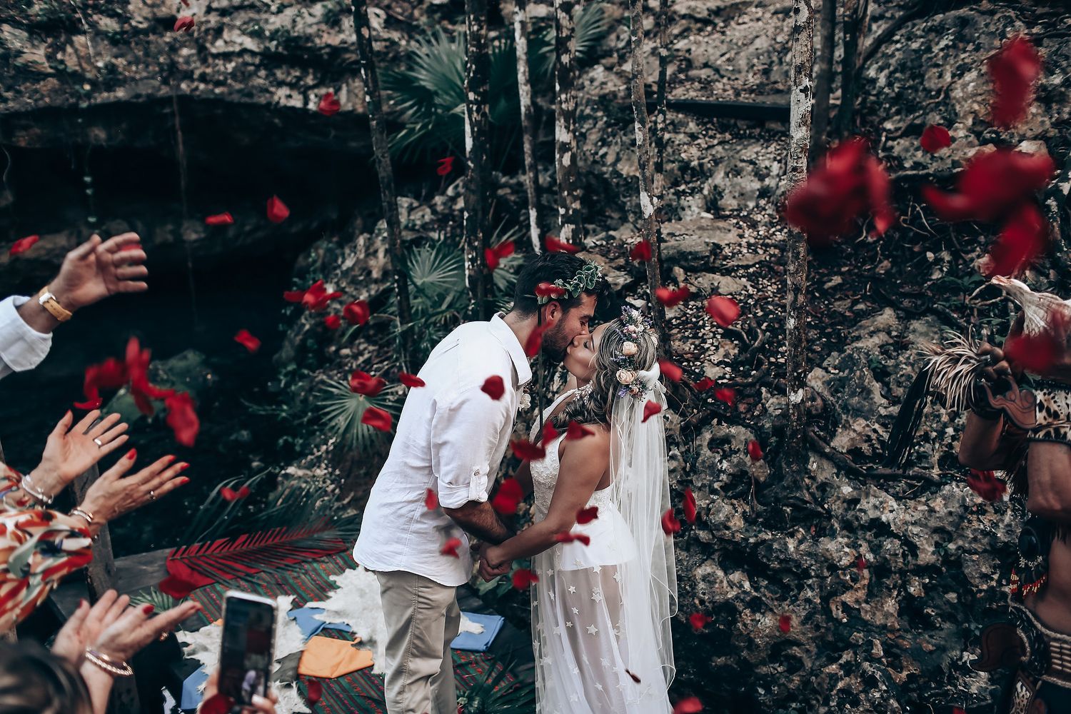 Participants in white attire celebrate outdoors with red elements visible in a natural forest setting.