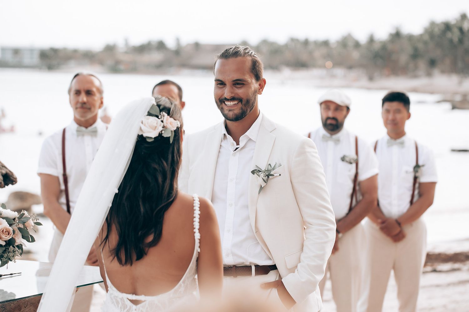 Wedding party members in white attire pose together on a tropical beach.