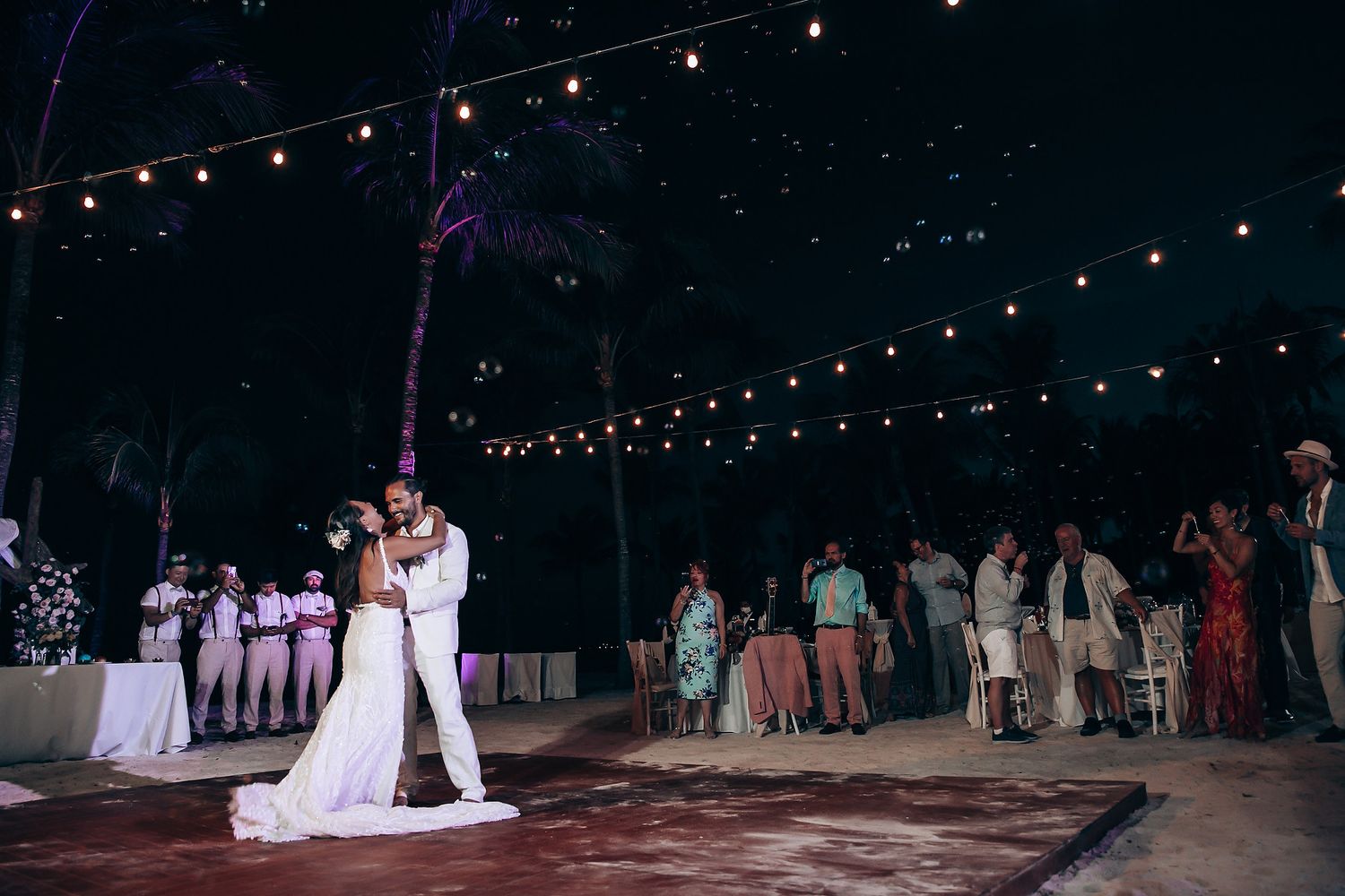 Newlyweds share their first dance under string lights at a nighttime beach reception.