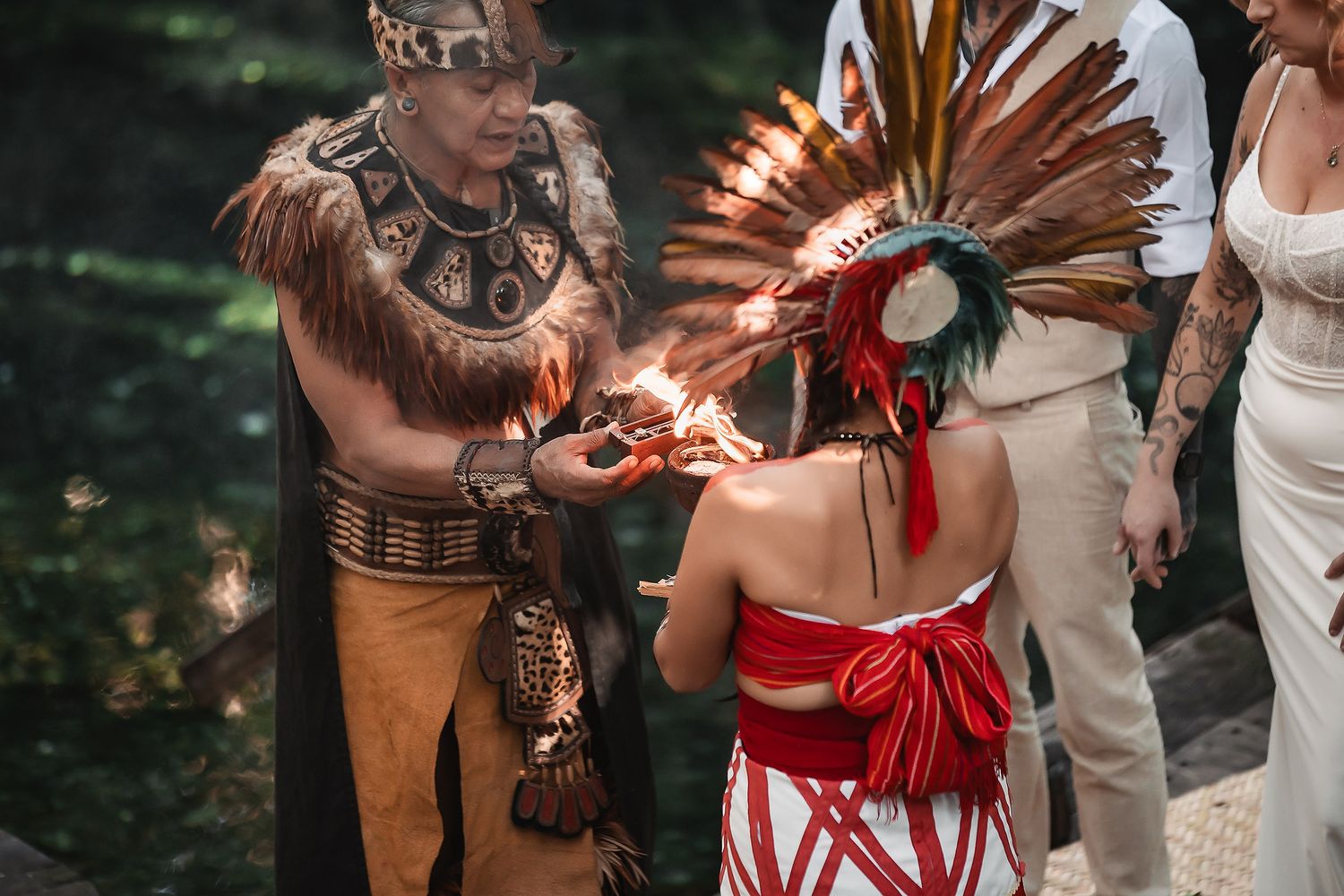 Two people in traditional Aztec ceremonial attire perform a ritual with fire during an outdoor cultural ceremony.