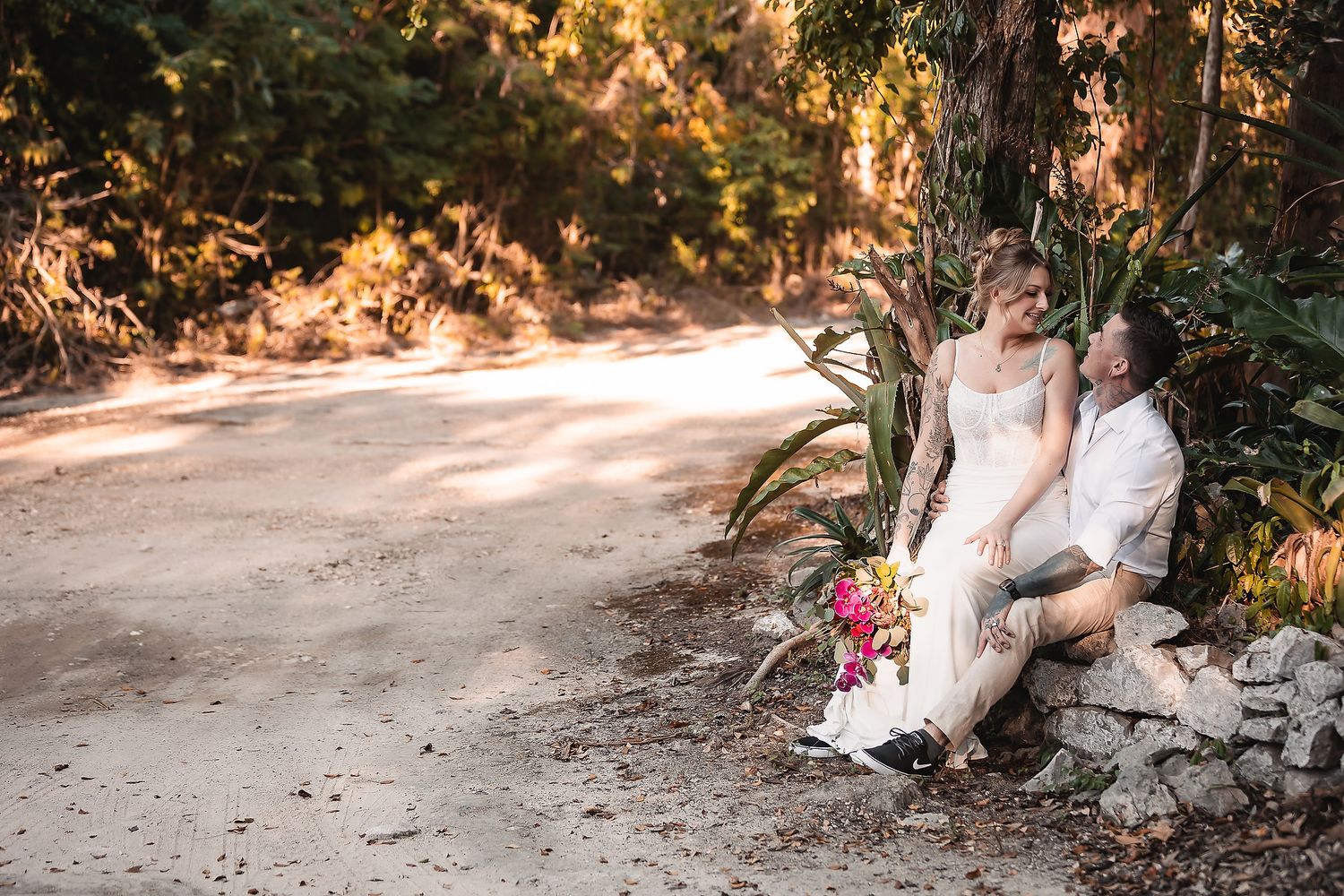 Bride and groom sitting together against rocks on a dirt path surrounded by lush tropical greenery on their wedding day.