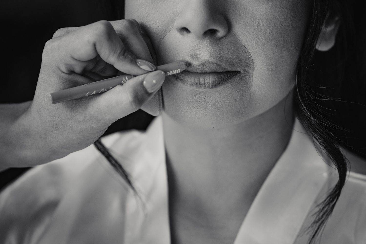 A makeup artist applies lipstick to a bride during wedding preparation in black and white photo.