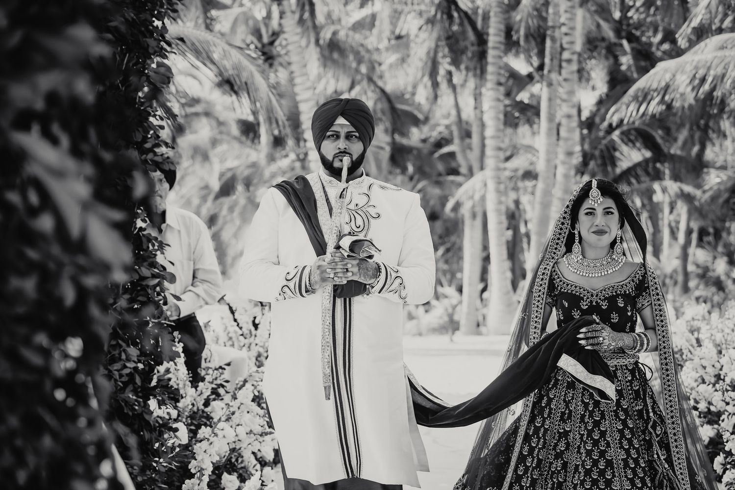Black and white photo of Sikh groom in turban walking with bride during traditional Indian wedding ceremony.