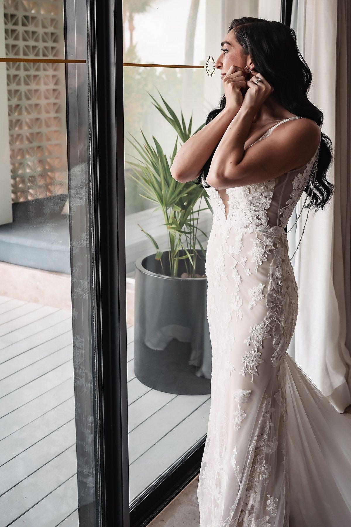 A bride in a fitted lace wedding gown puts on earrings while standing by a glass door with natural light.