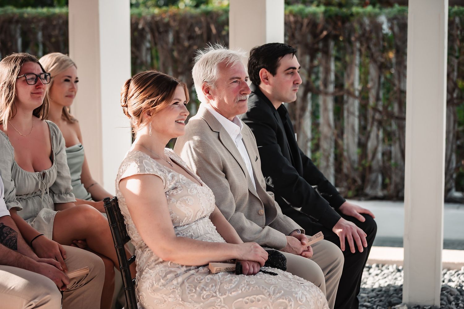 Wedding guests smiling and seated during an outdoor ceremony, dressed in elegant attire including lace and suits.