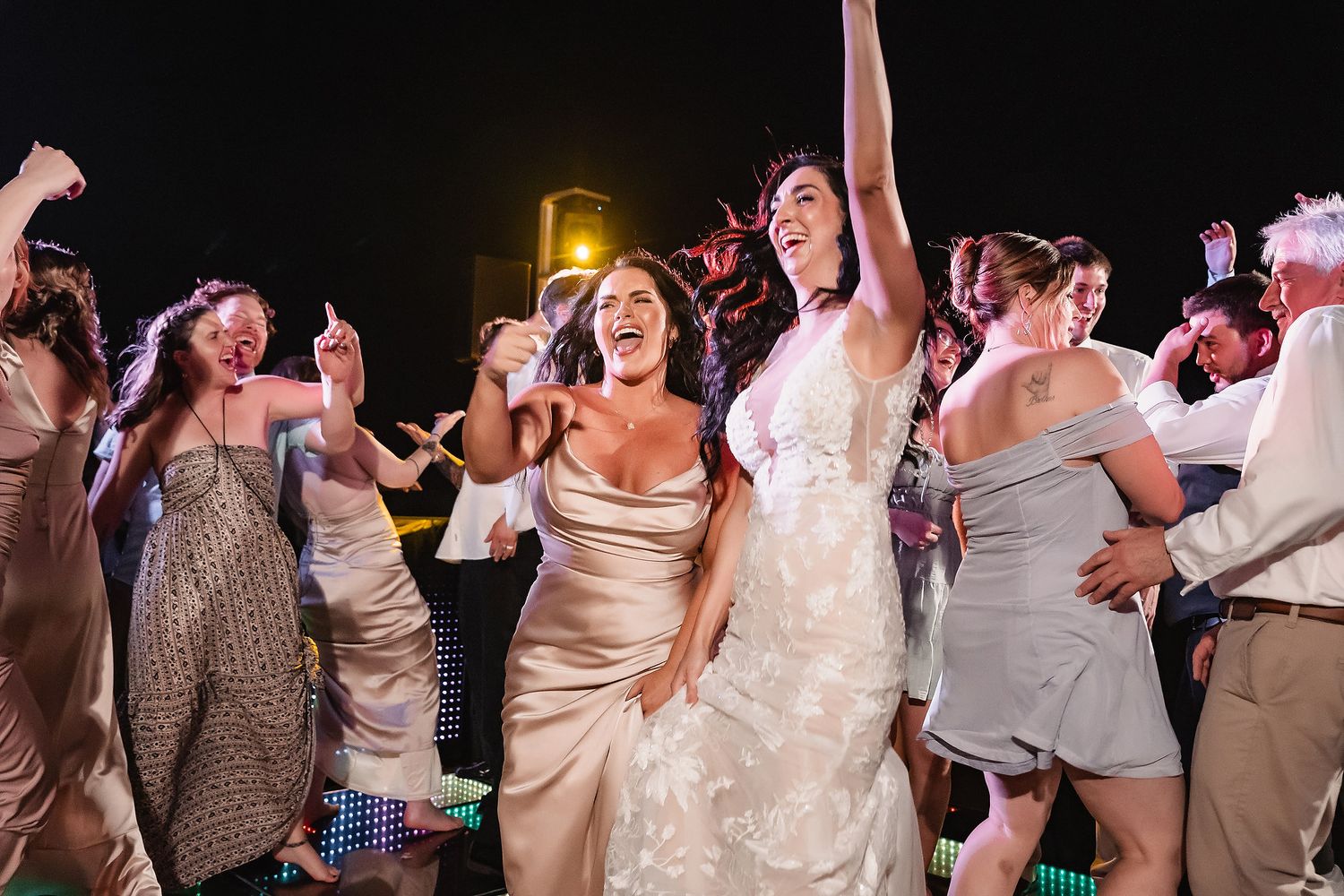 Bride in white gown dancing joyfully with guests at outdoor wedding reception at night.