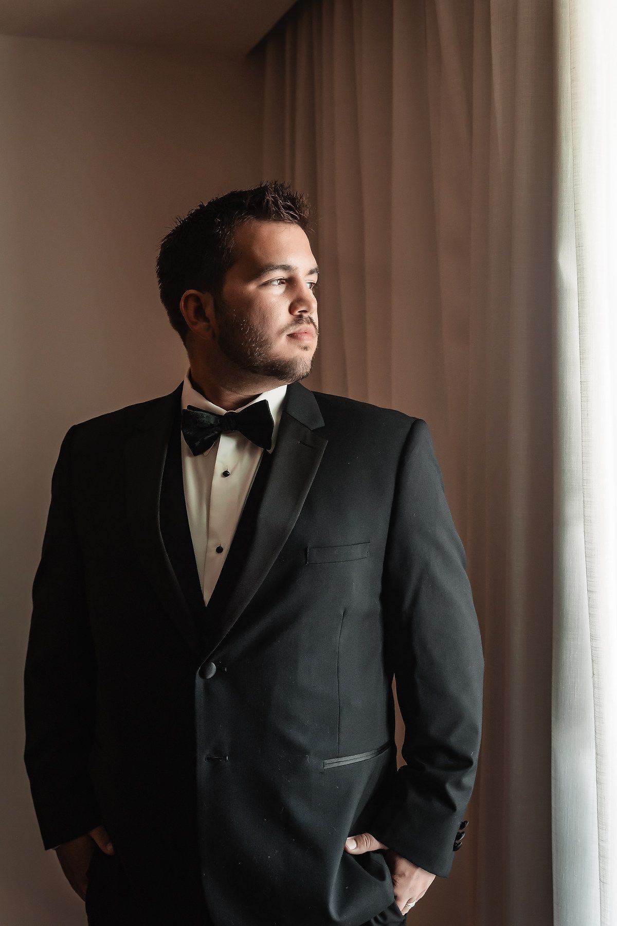 A well-dressed man in a black tuxedo with bow tie stands by a curtained window, gazing thoughtfully to the side.