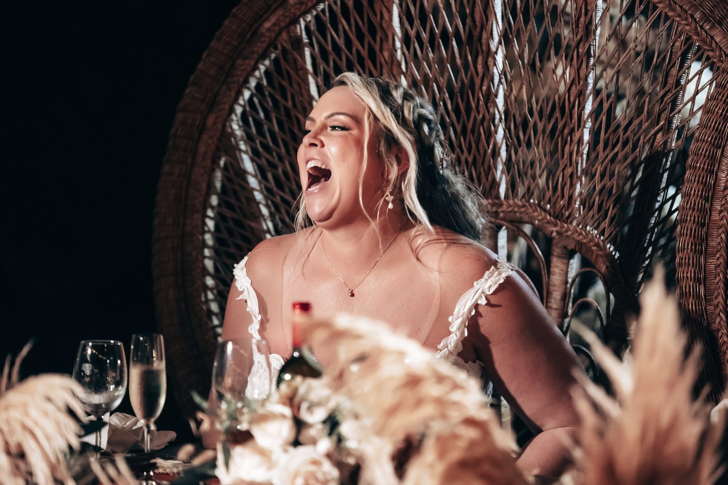 Joyful blonde bride laughing at wedding reception table decorated with pampas grass.