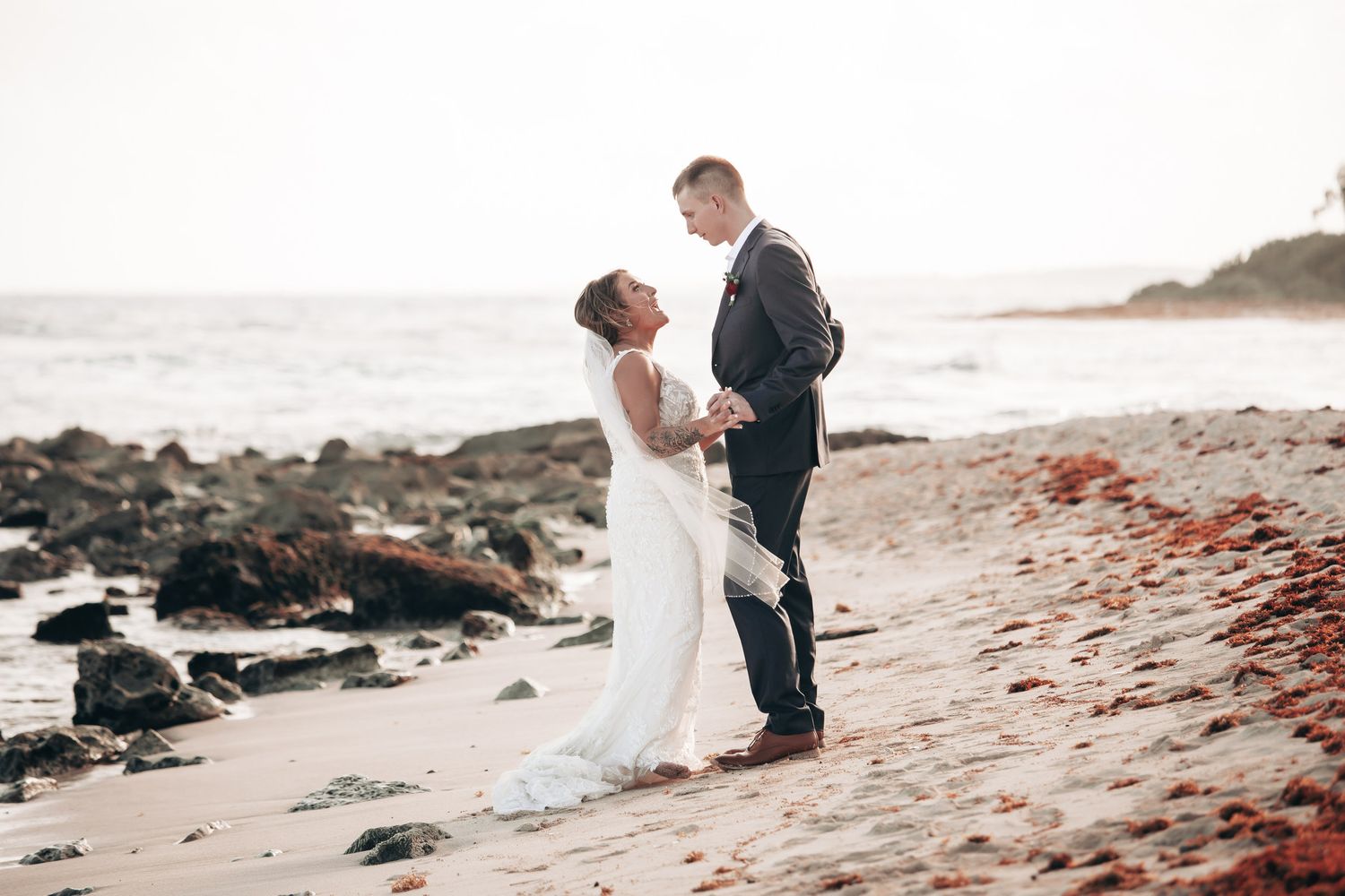 Bride and groom share a tender moment holding hands on a scenic rocky ocean beach.