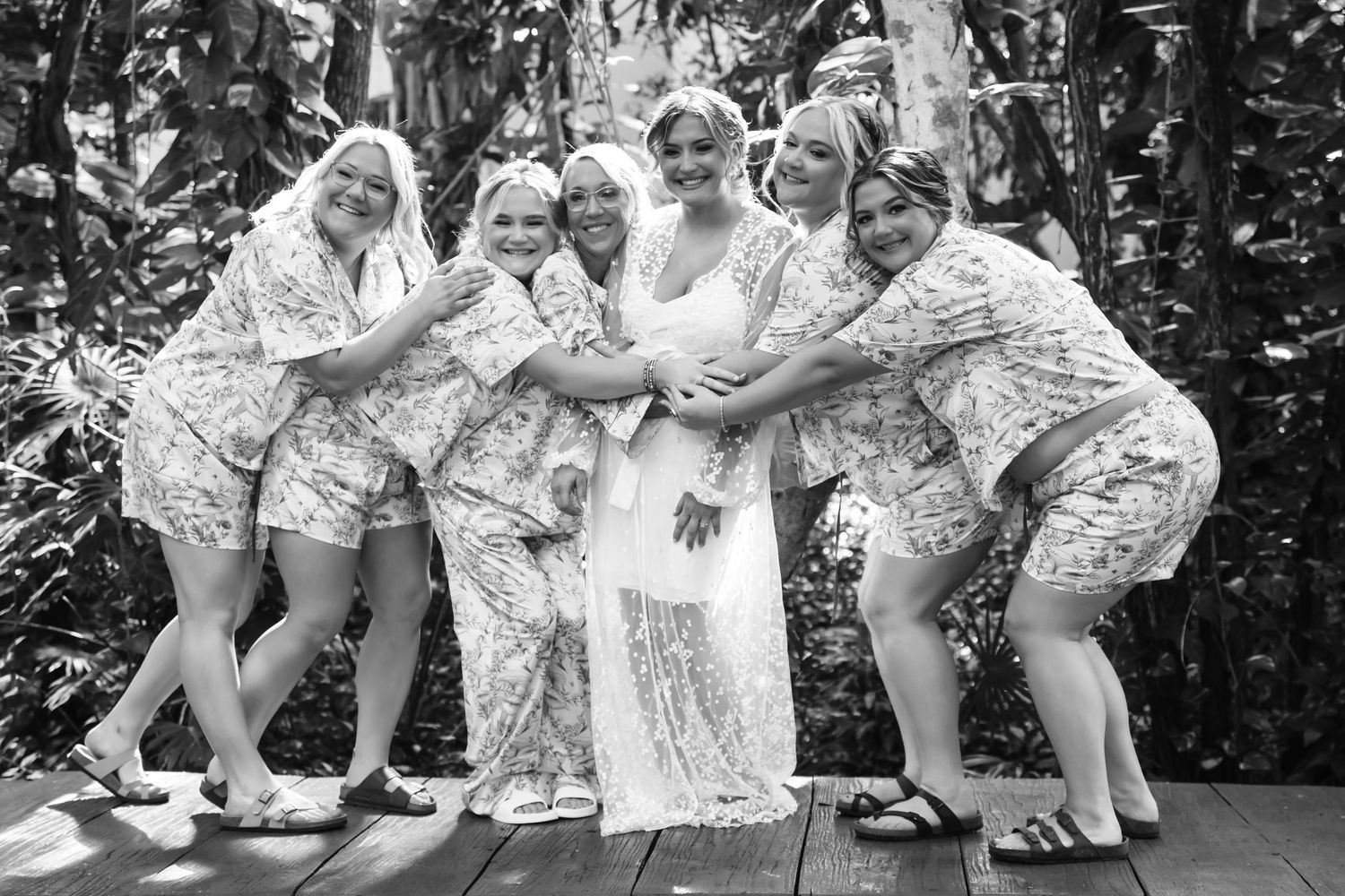 Bride and bridesmaids in matching floral robes posing together on tropical wooden deck.