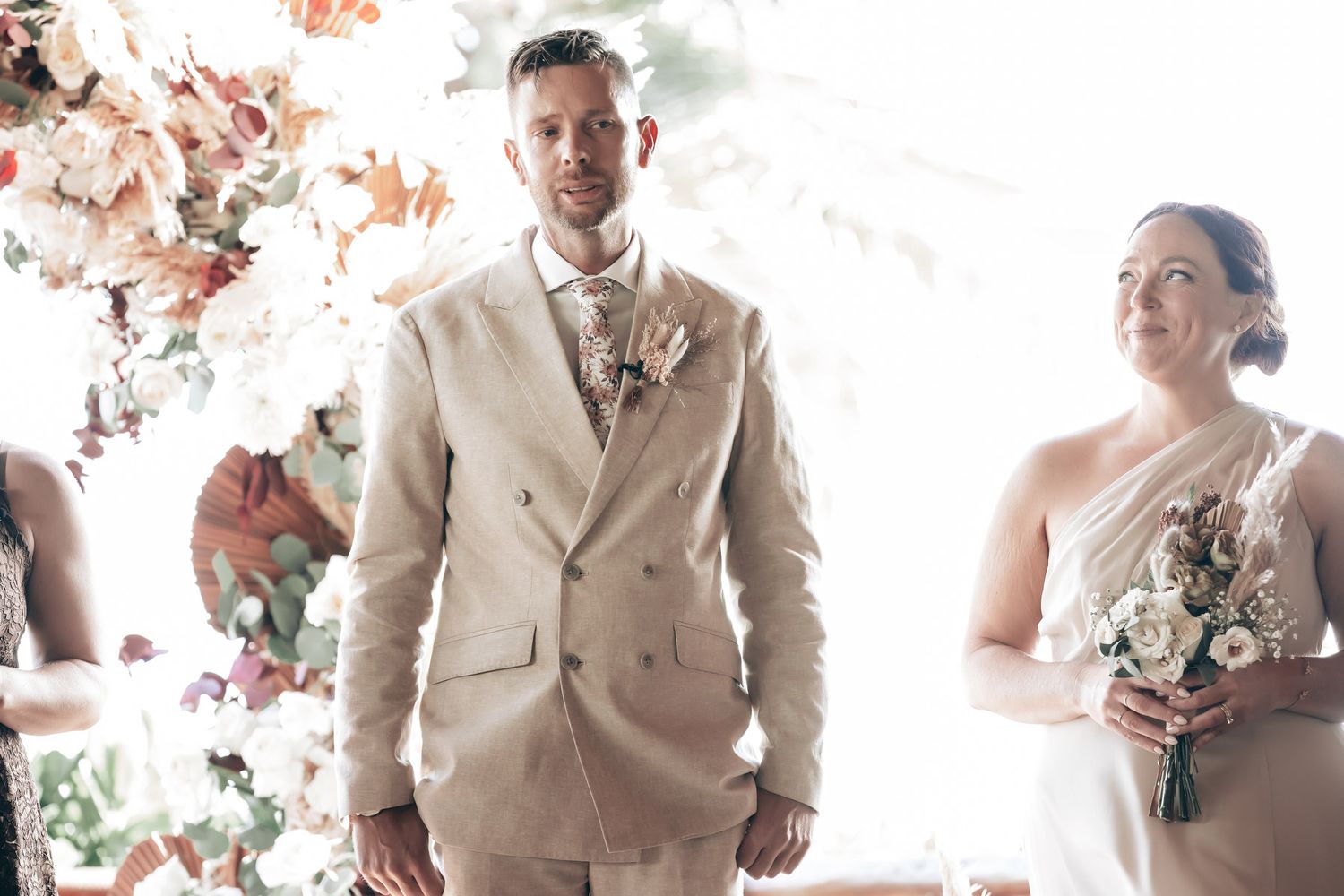 Groom in beige double-breasted suit standing at the altar with a floral boutonniere during wedding ceremony.