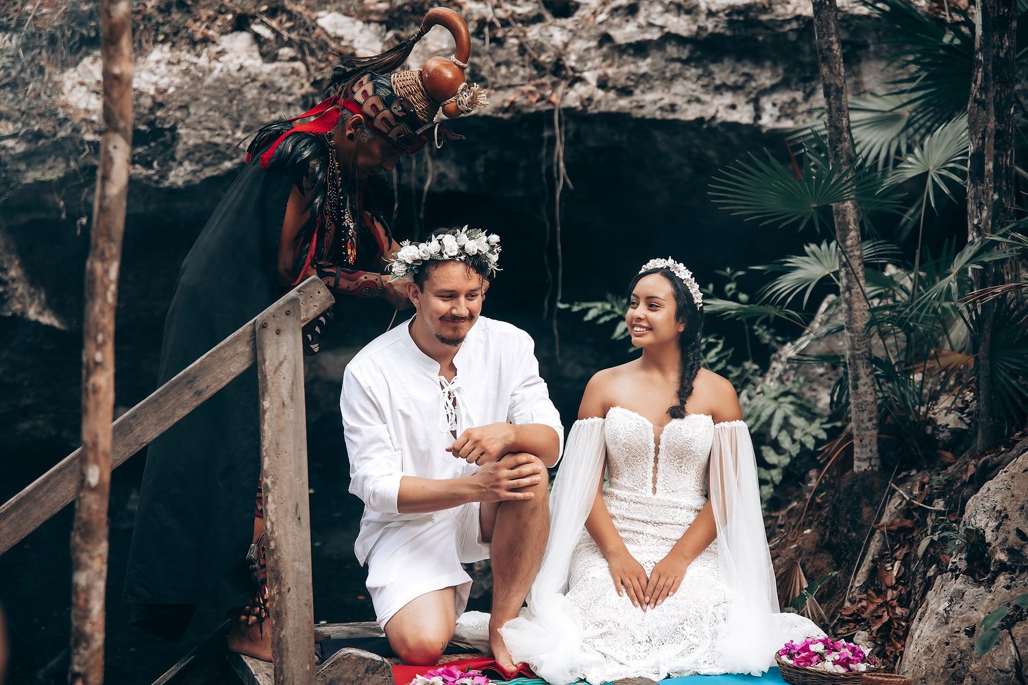 A couple in white attire sits together on a stone wall in a tropical garden setting.