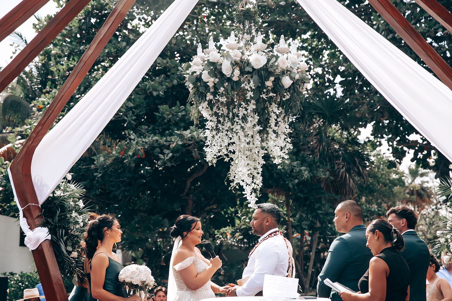 Outdoor wedding ceremony under wooden arch with flowing white fabric and natural greenery.
