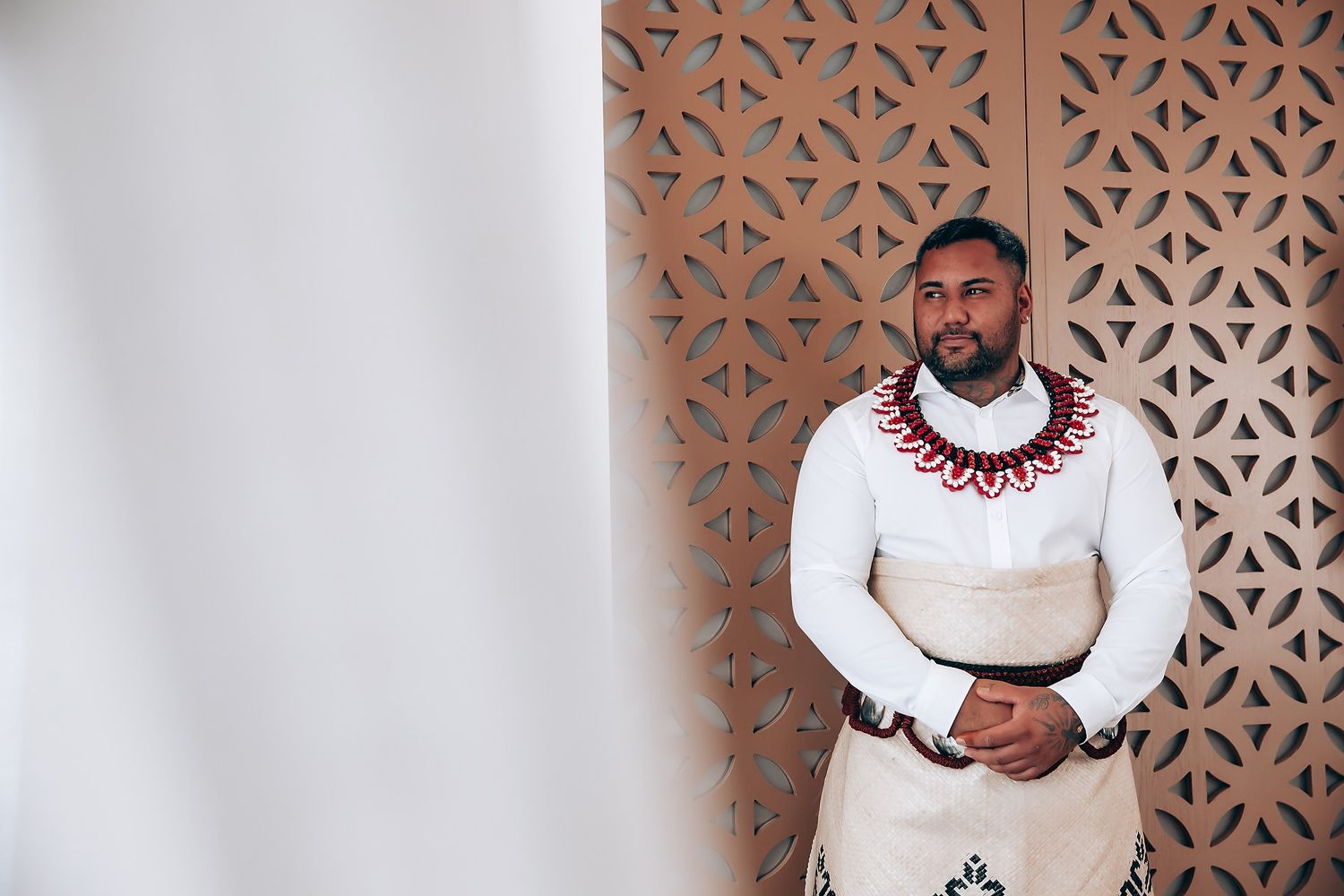 A person wearing a white dress shirt with a red beaded necklace stands against a decorative lattice wall.