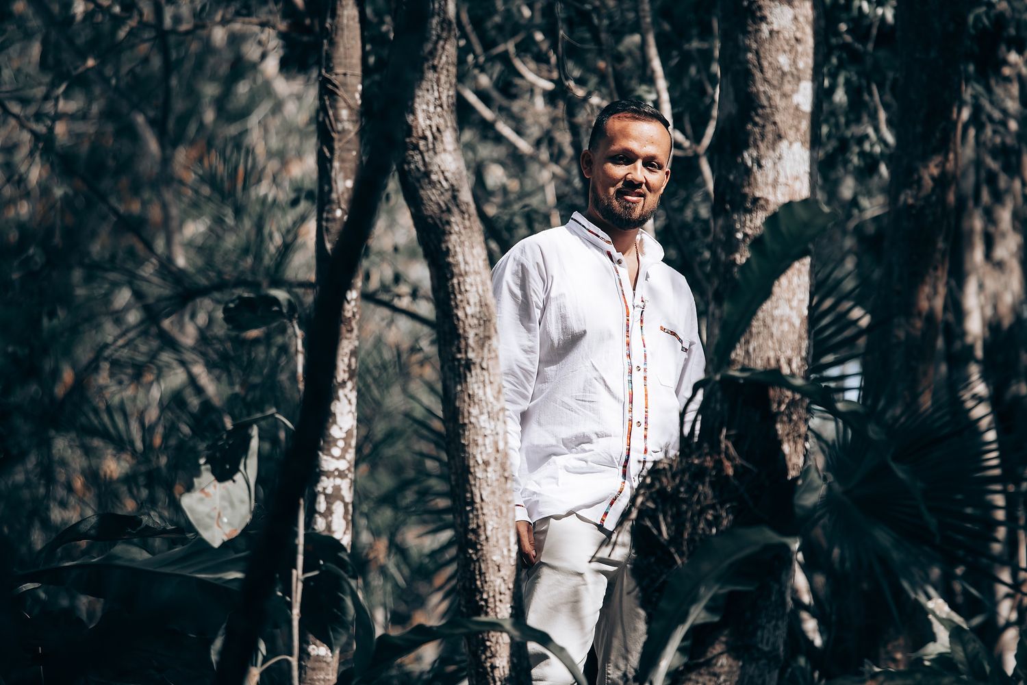 A person in a white shirt and light pants smiles while standing in a forest setting with sunlight filtering through trees.