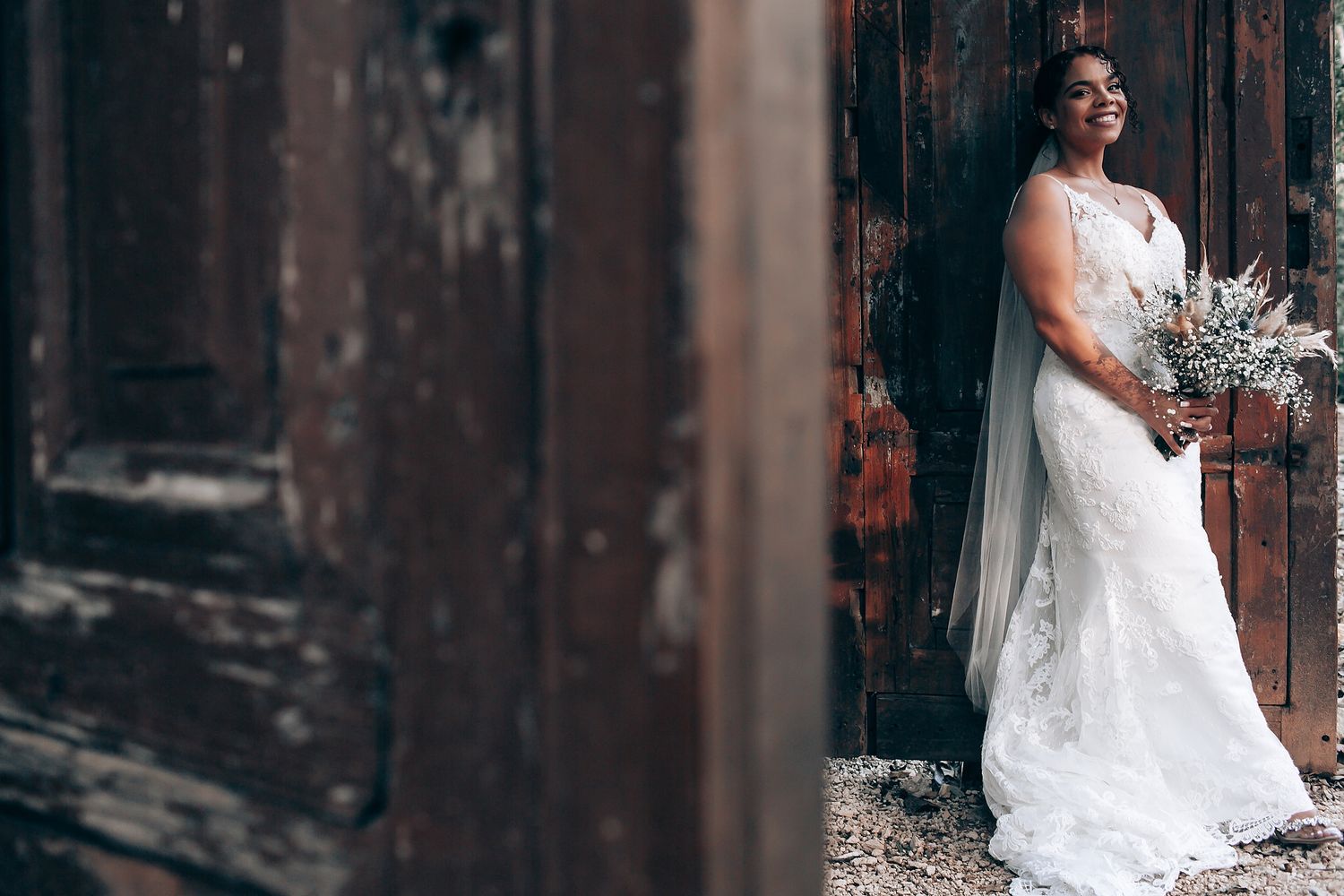 Bride in white lace dress poses against dark wooden backdrop.