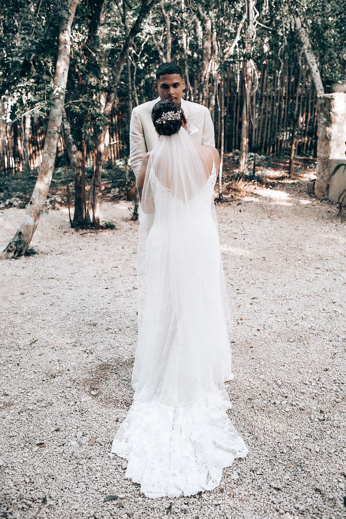 A flowing white wedding dress photographed from behind in a wooded outdoor setting.