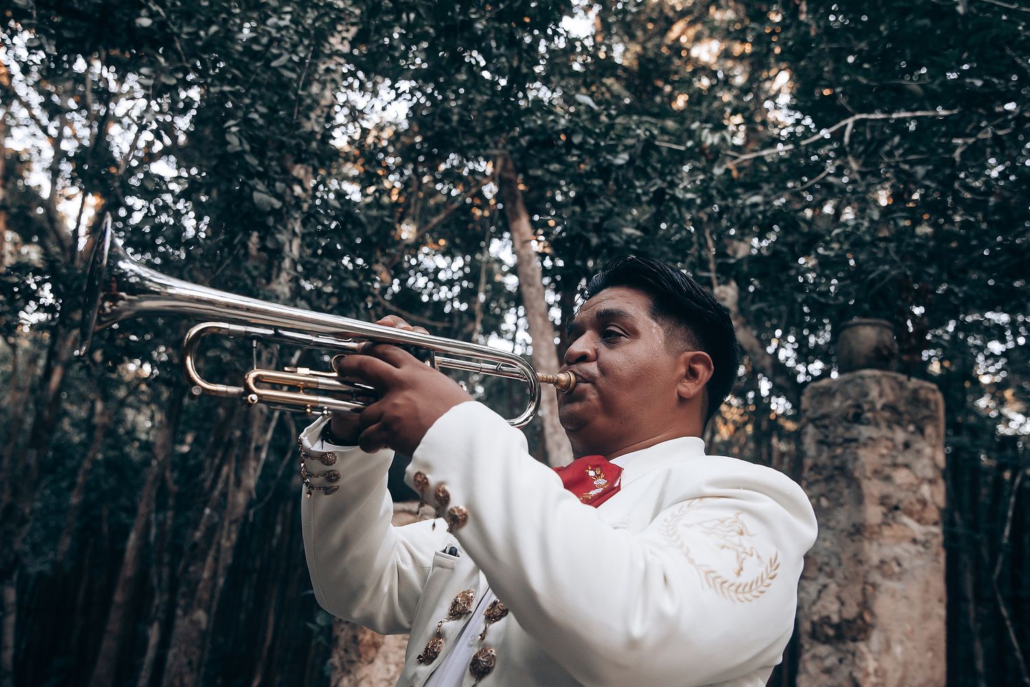 A musician in white formal attire plays trumpet during an outdoor forest performance.
