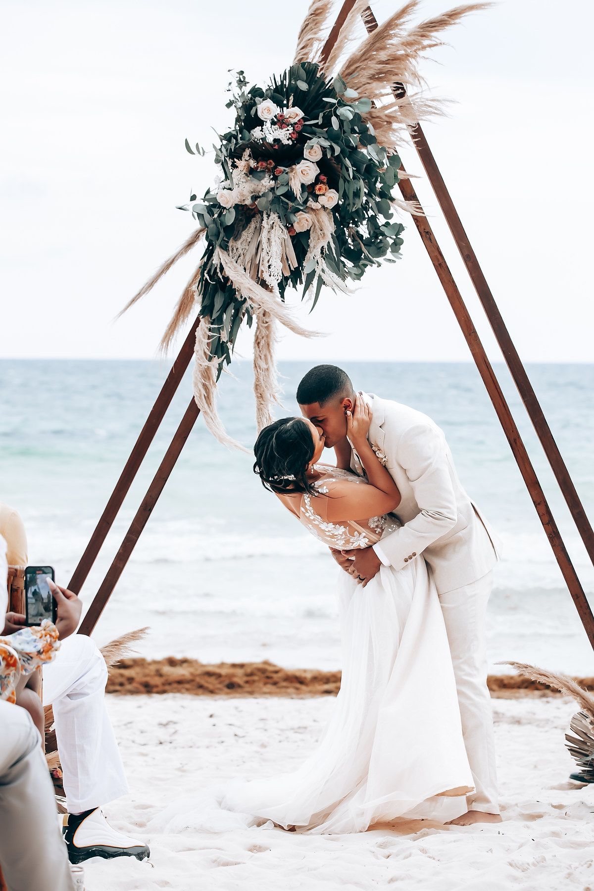 Romantic beach wedding ceremony under wooden arch decorated with pampas grass and greenery by the ocean.