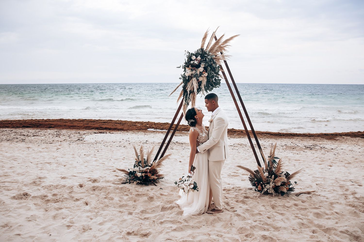Bohemian beach wedding setup with wooden arch decorated with tropical flowers and greenery.
