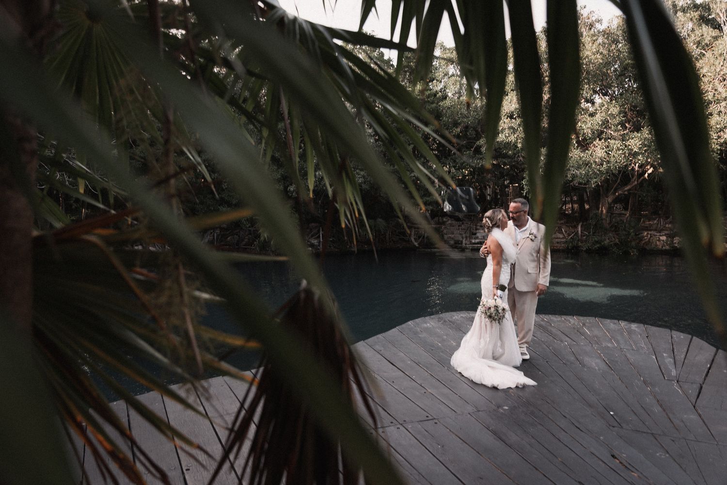 Bride and groom standing on wooden deck surrounded by tropical palm leaves near serene blue water.