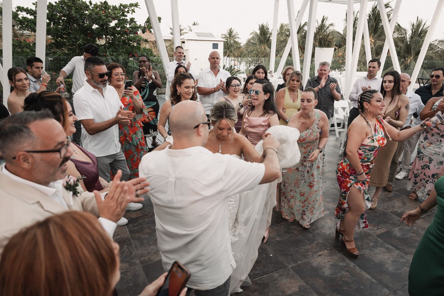 Newlyweds celebrate joyfully with cheering wedding guests at an outdoor tropical reception venue.