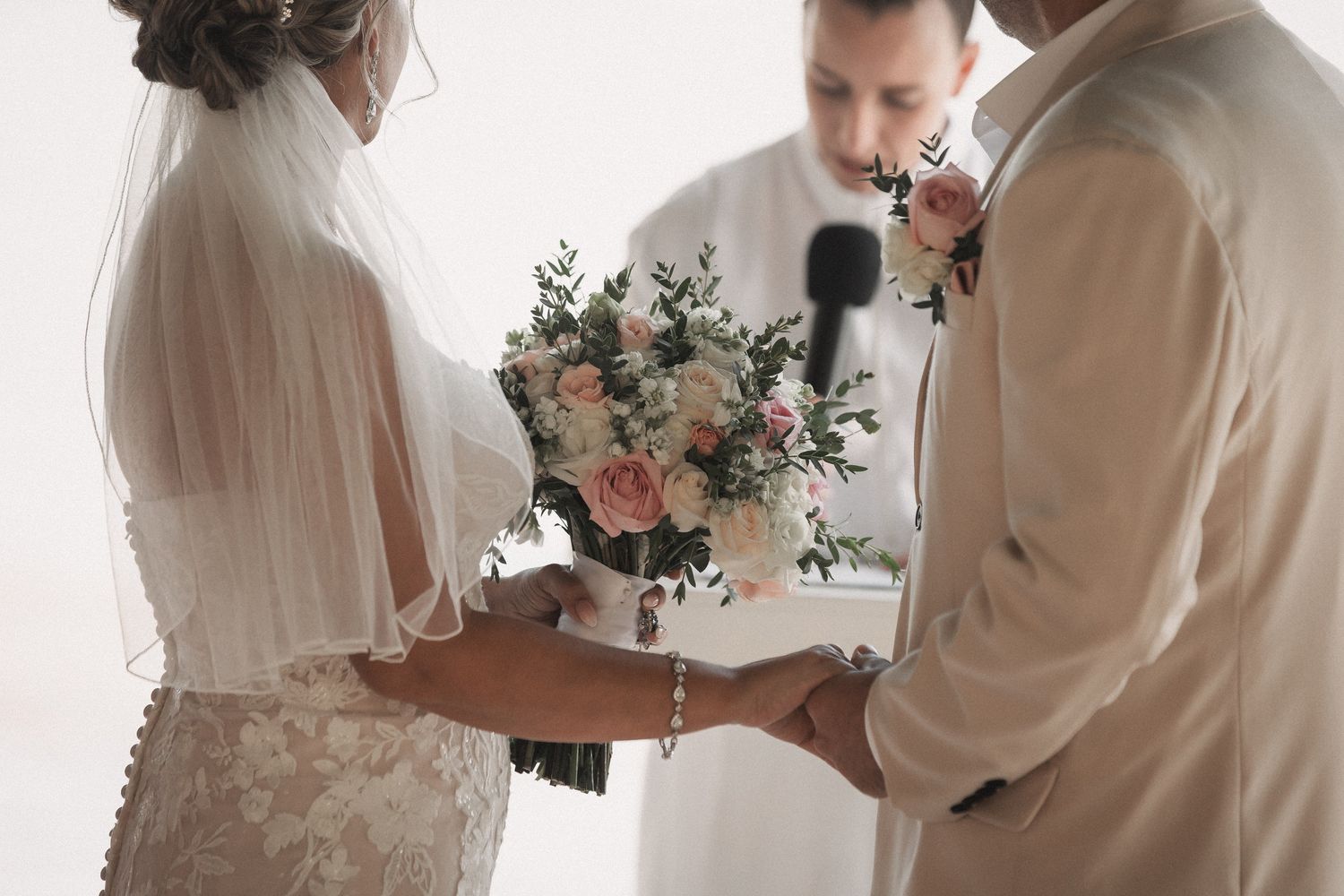 Bride and groom hold hands during wedding ceremony while officiant reads vows, bride holding pink rose bouquet.