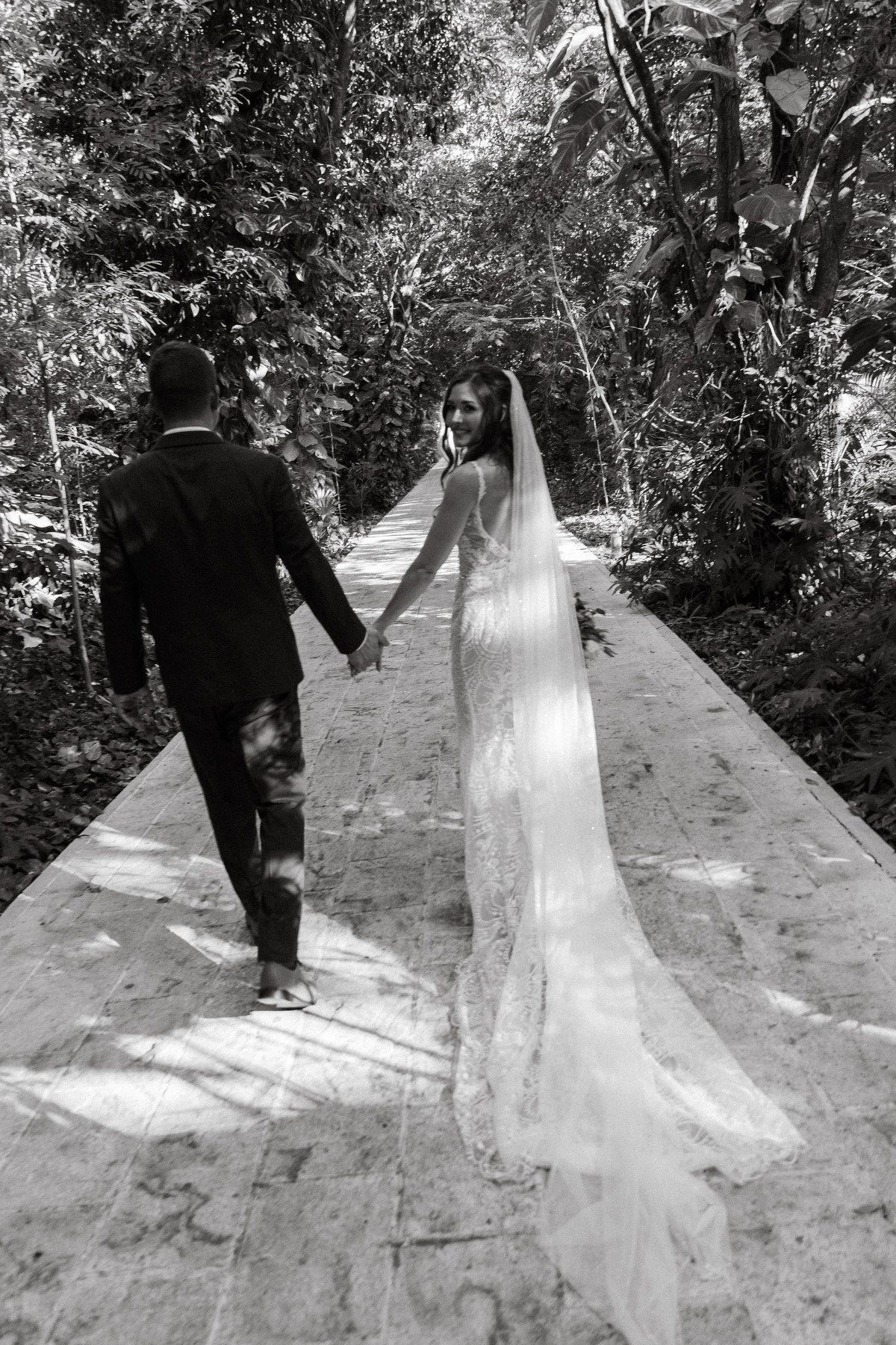Black and white photo of bride and groom holding hands walking through tropical path.