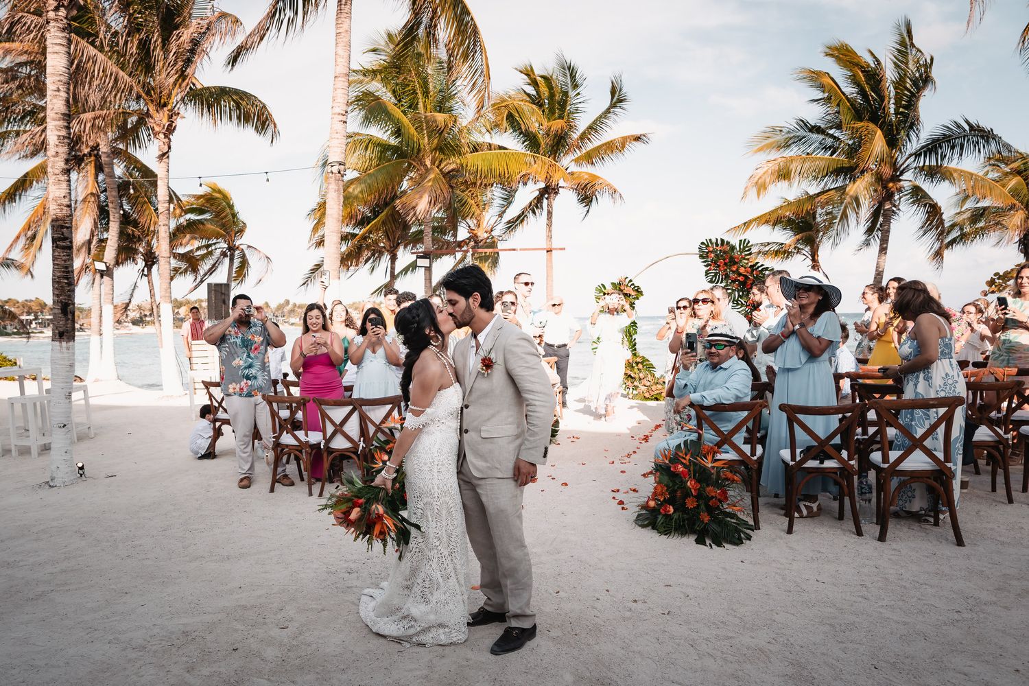Newlyweds share first kiss on sandy beach aisle surrounded by palm trees and guests at tropical destination wedding.