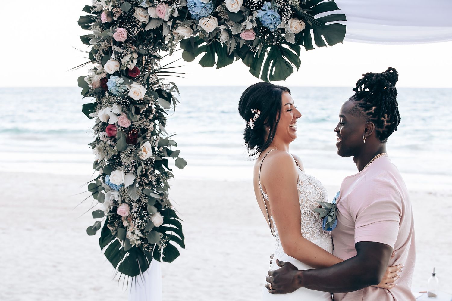 A close-up of a bride and groom embracing in front of a floral arch on a beach.