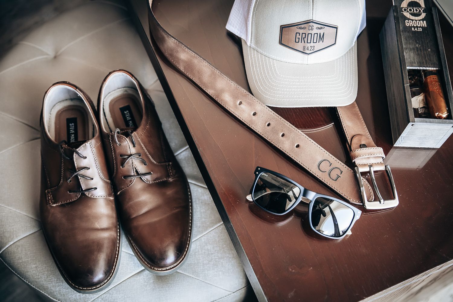 Leather dress shoes, a brown leather belt, and sunglasses on a wooden surface.
