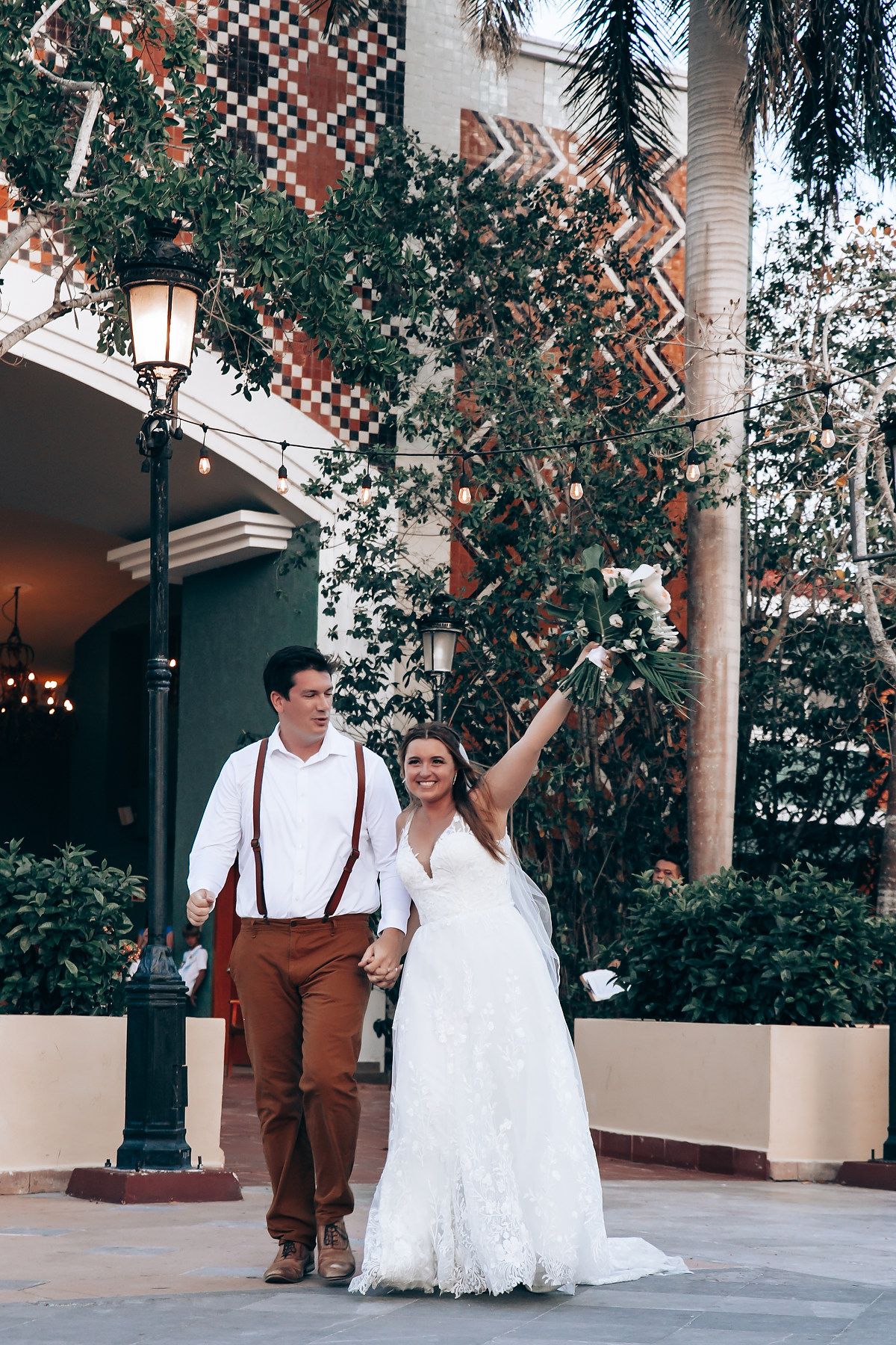 Couple walks together under palm trees and string lights at tropical wedding venue.
