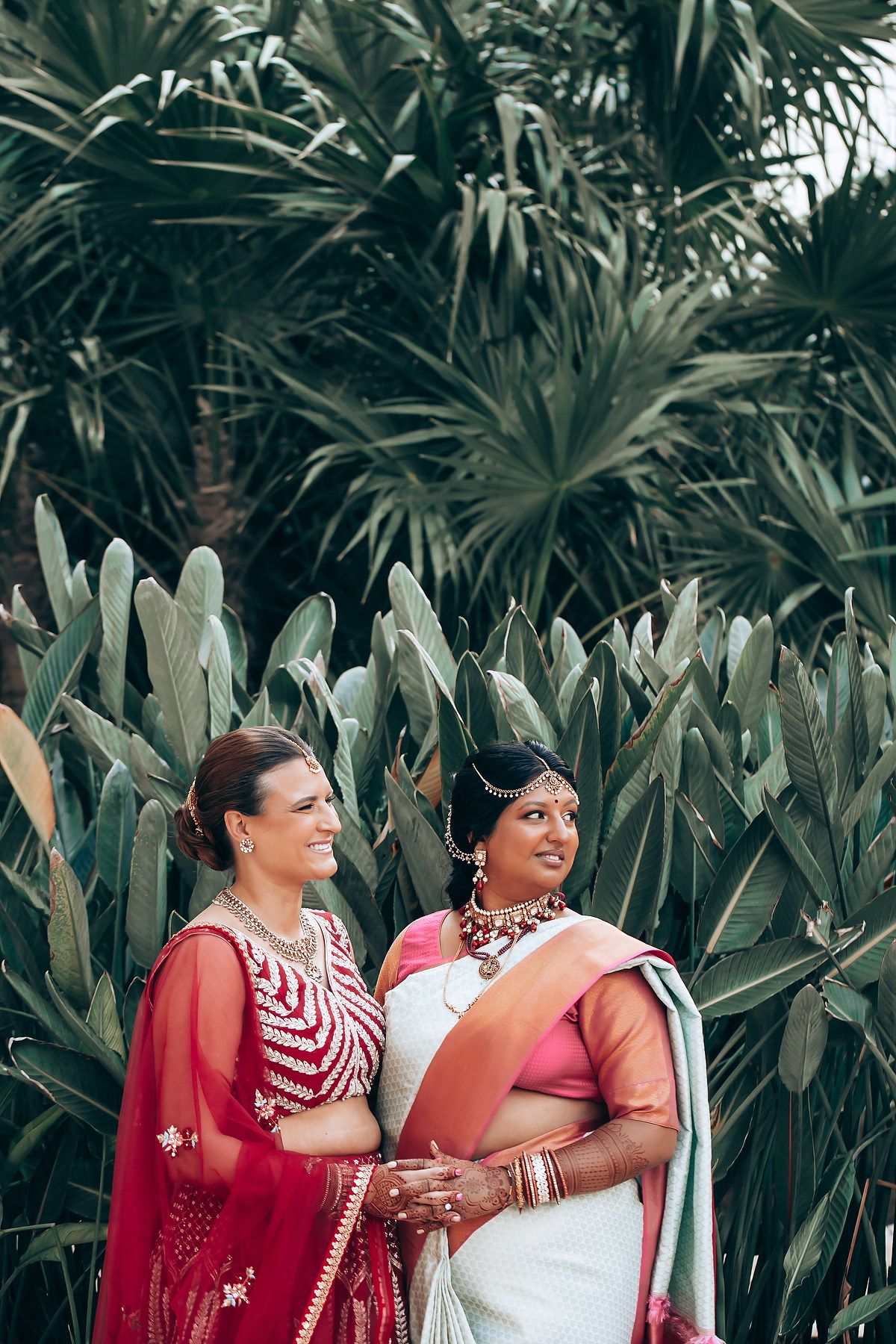 Two women in traditional red Indian wedding attire share a joyful moment against a lush tropical backdrop.