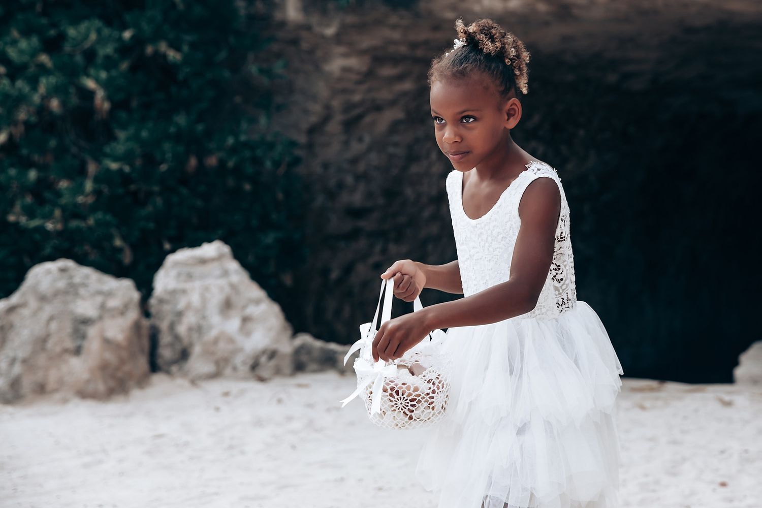 Young flower girl in a white tulle dress sitting on a sandy beach near rocks.