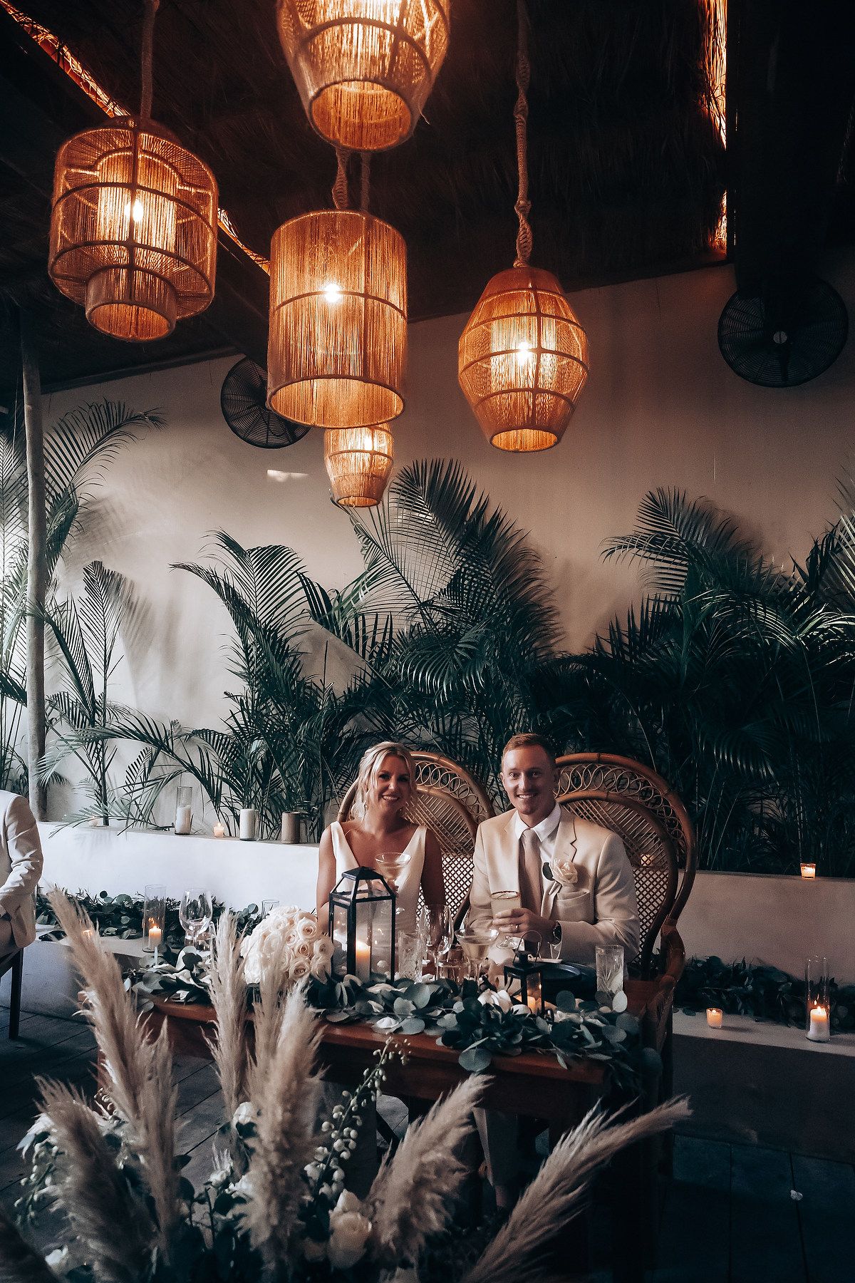 Elegant reception setup with rattan pendant lights and tropical palm decor creating a warm, moody ambiance.