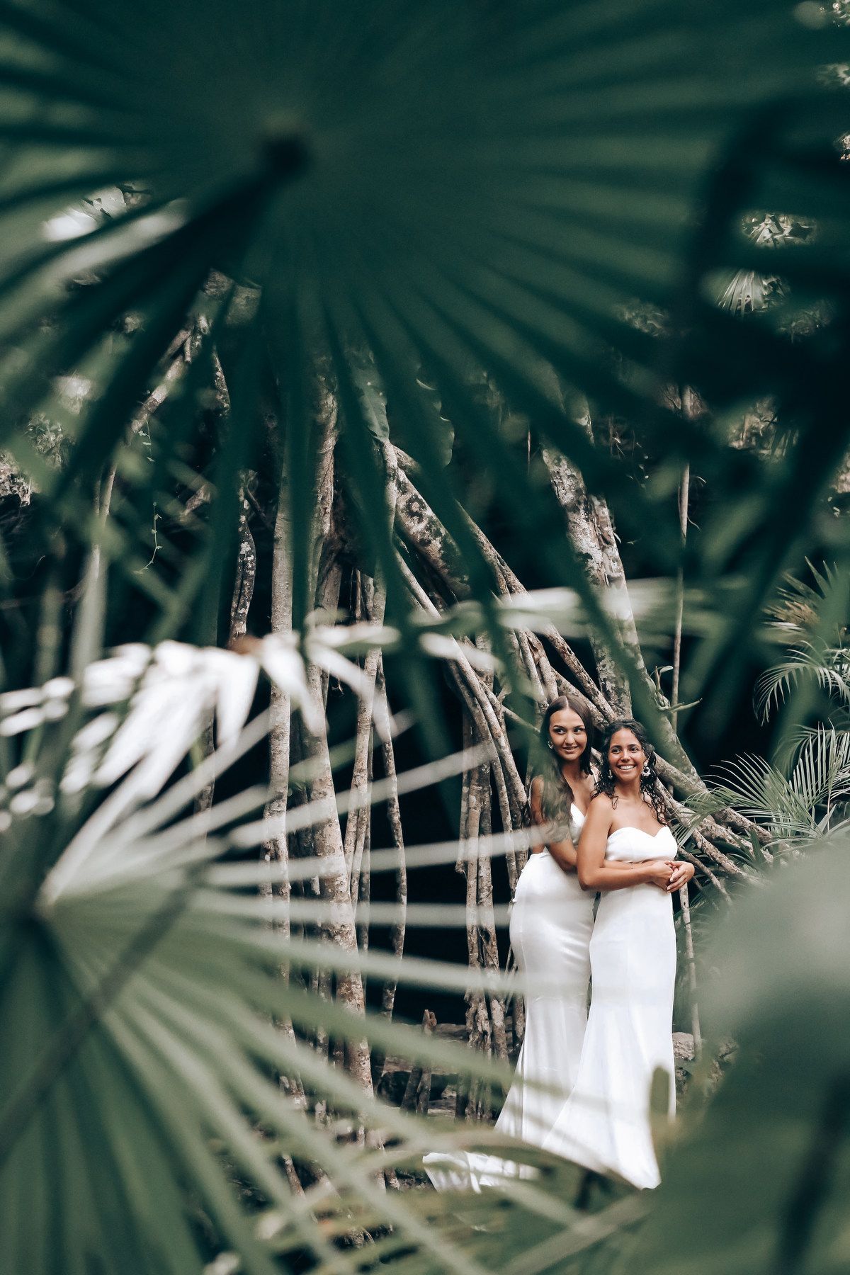 Couple in white dresses framed by tropical palm fronds in a natural setting.