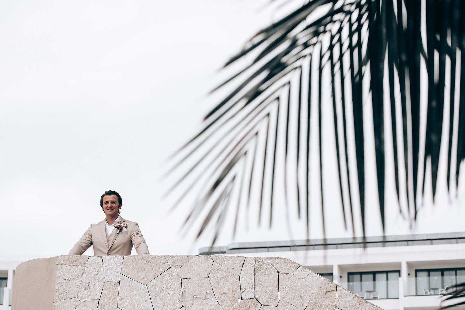 Person in light colored clothing standing behind a stone wall with palm fronds visible above.