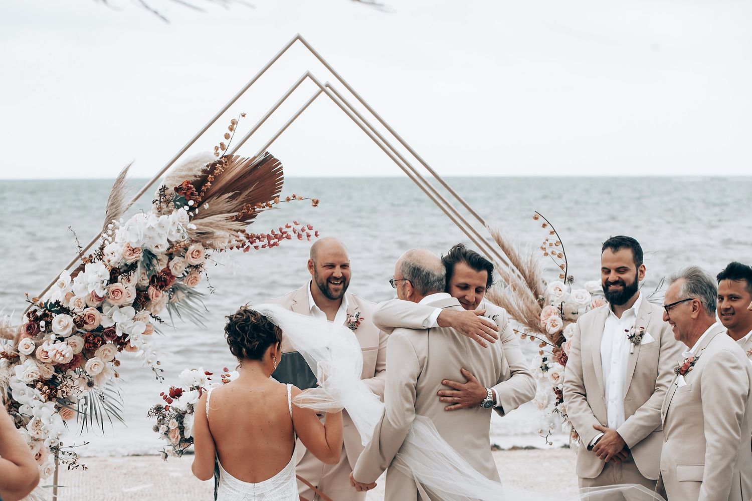 Geometric wedding arch decorated with flowers frames guests at beachside ceremony.
