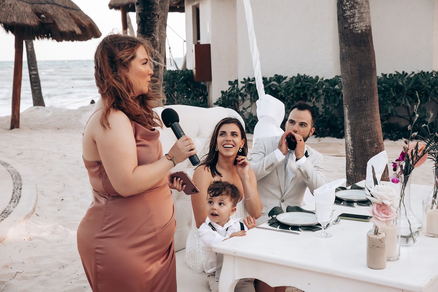 Wedding guests enjoying a reception at tables near the beach with tropical palm trees.