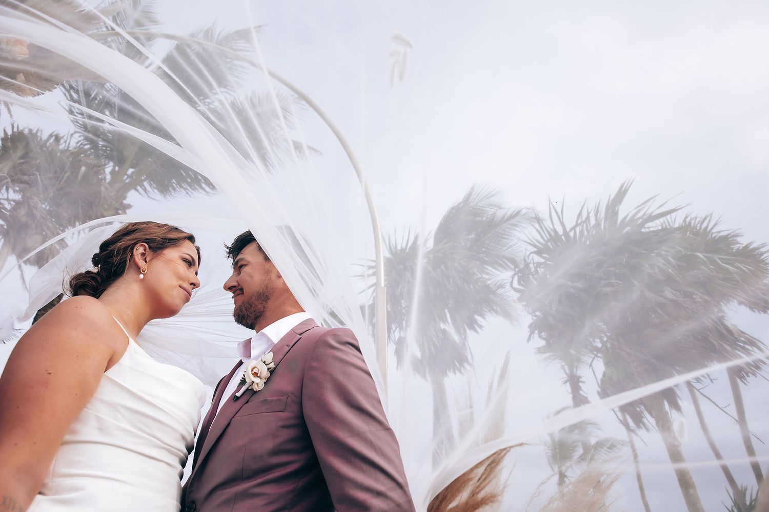 A flowing white wedding dress catches the breeze on a palm-lined beach.