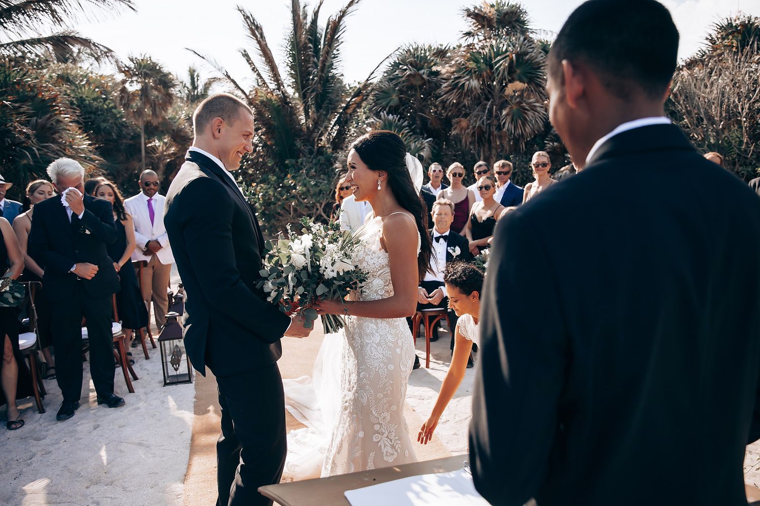 Outdoor wedding ceremony with guests gathered under palm trees.