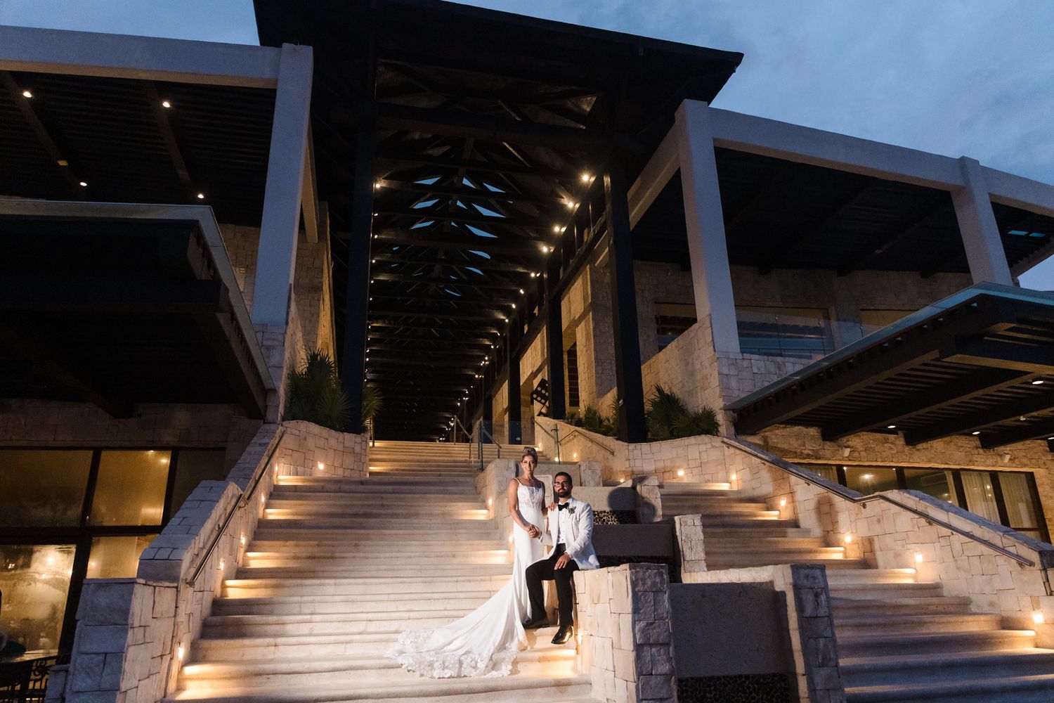 Bride and groom pose on illuminated stone steps of elegant venue at dusk in romantic wedding portrait.