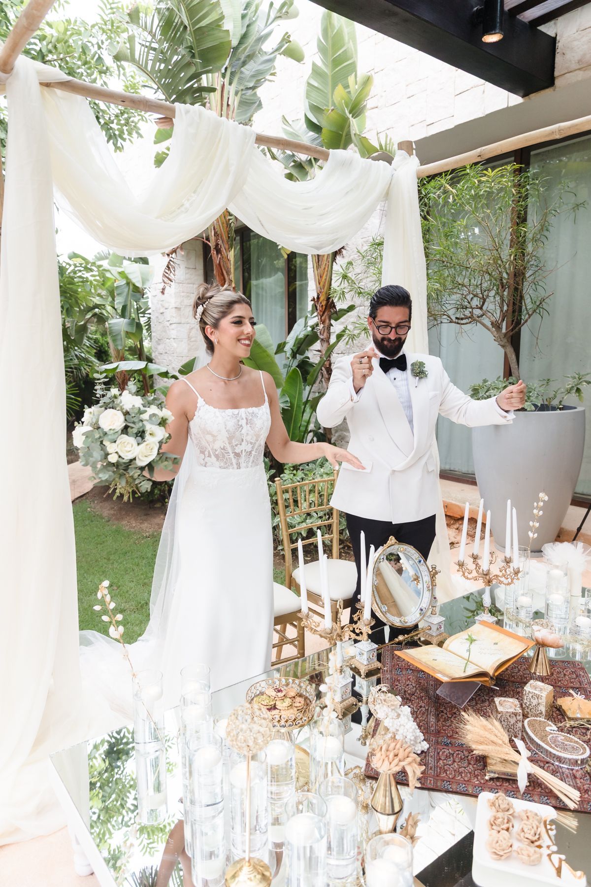 Bride and groom in white attire stand beneath draped fabric canopy at elegant outdoor wedding reception table.