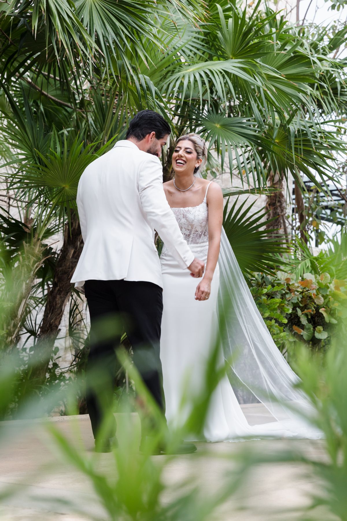 Bride and groom holding hands and smiling at each other outdoors, surrounded by lush tropical palm trees.