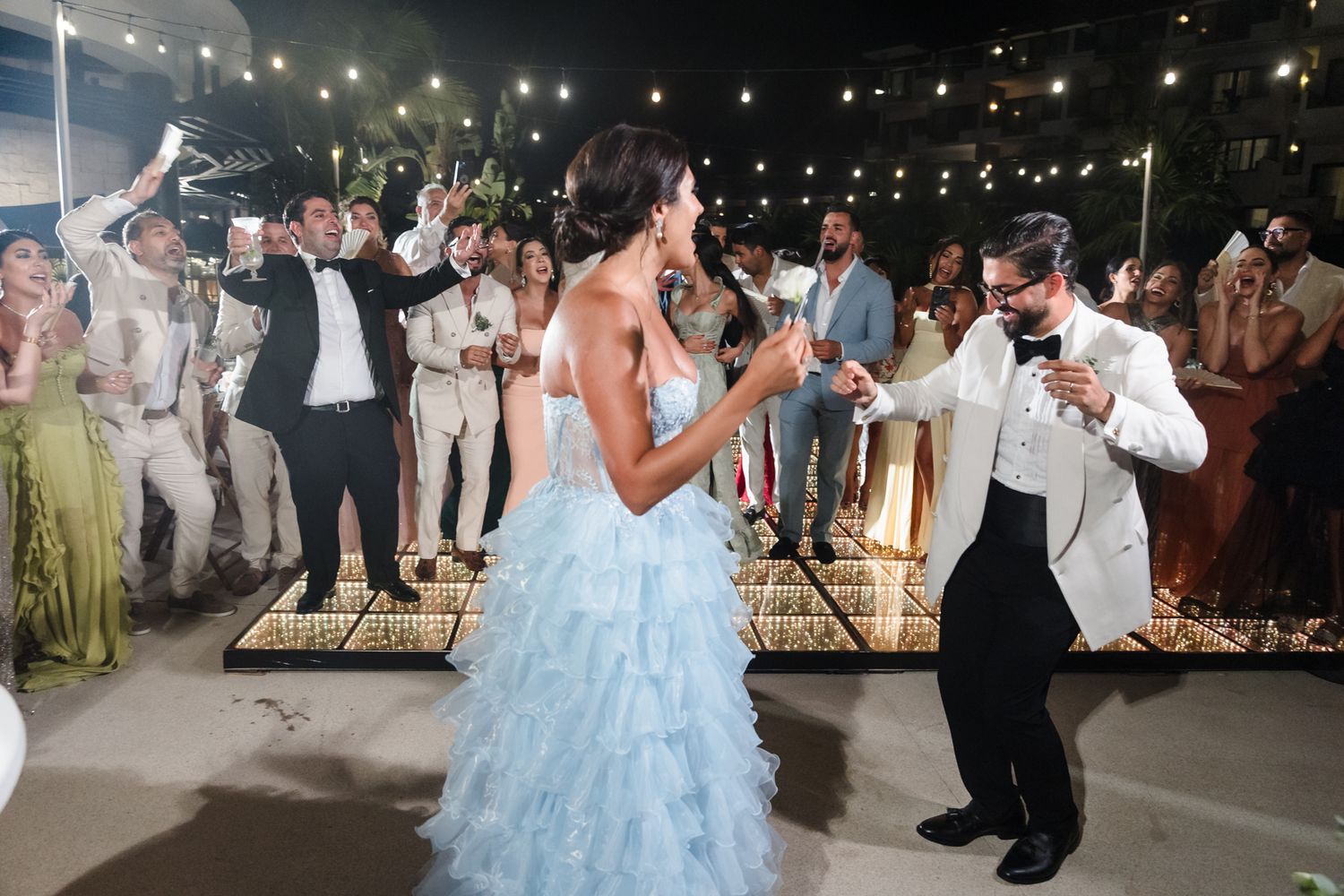 Couple shares first dance in blue ruffled gown at elegant outdoor wedding reception with guests celebrating.