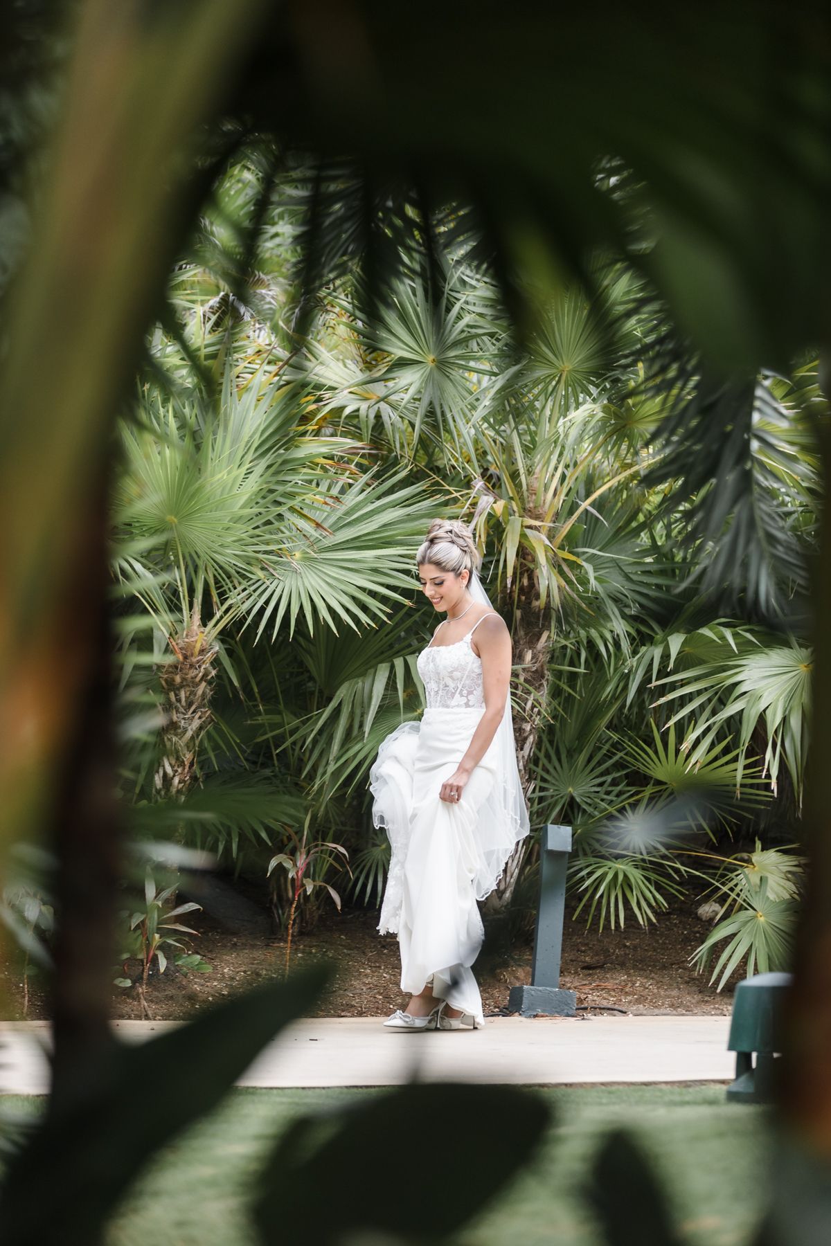 Bride in white gown walking among lush tropical palm trees in an outdoor garden setting.