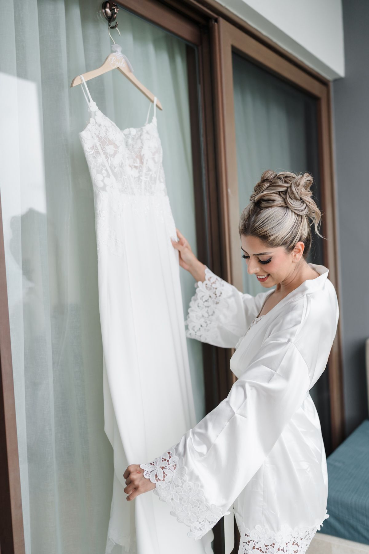 Smiling bride in white lace robe admiring her elegant wedding dress hanging by a bright window.