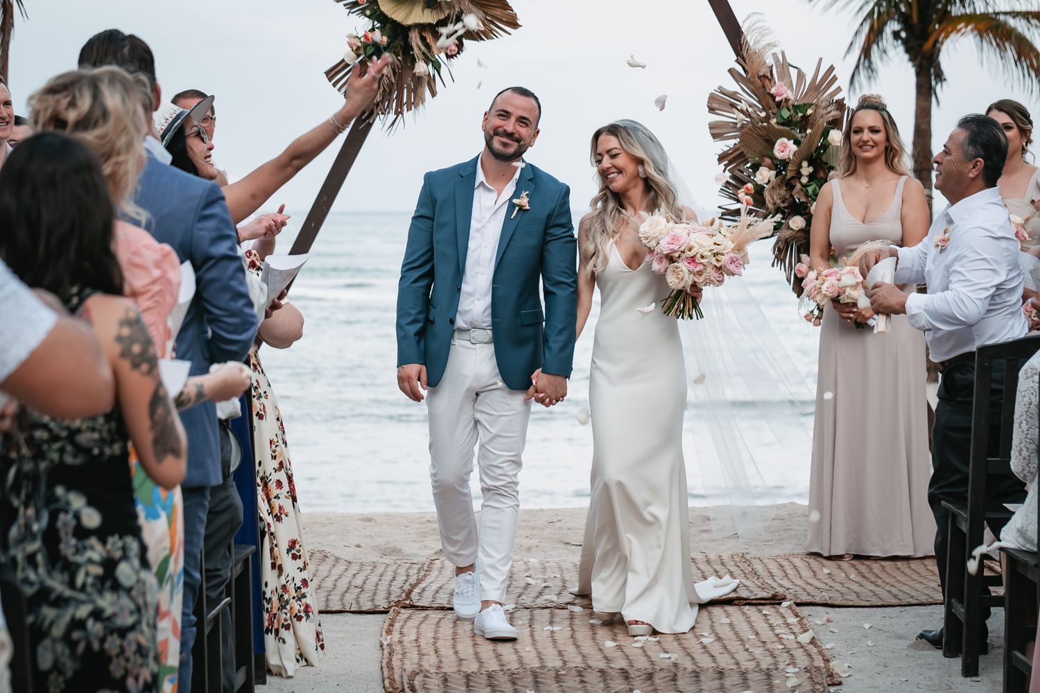 Newlyweds walk down the aisle at a beachside wedding celebration with guests cheering.